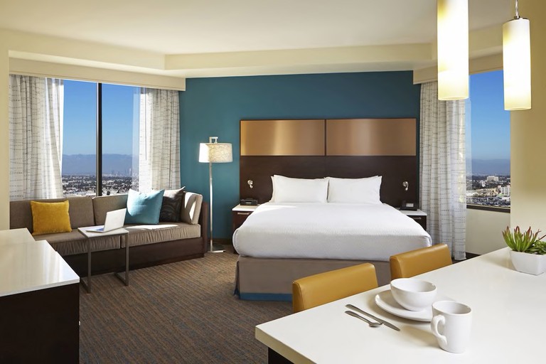 Corner room with large bed, tan sofa, dining table and city views at Residence Inn by Marriott Los Angeles LAX/Century Boulevard